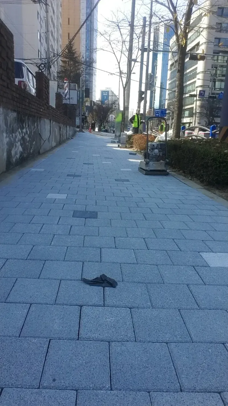 강남 경기고 사거리 인근 보도 시공 중 모습. 연회색 정사각 블록이 새로 깔린 보도가 담장을 따라 뻗어 있고 앞쪽 바닥에 장갑이, 멀리 다짐기와 작업자가 보인다.