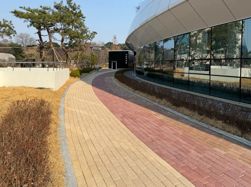 올림픽회관 유리 외벽을 따라 휘어지는 보행로. 베이지 블록 길 가장자리에 적갈색 블록 띠가 곡선으로 이어진다.