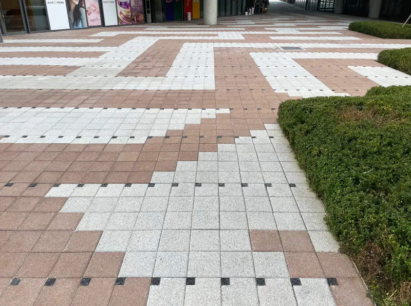 중곡 보건복지행정타운 광장 바닥. 적갈색 블록 사이로 밝은 회색 블록 띠가 굽이치듯 꺾이고 오른쪽에 낮은 회양목 화단이 있다.