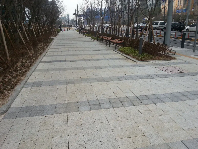 판교 SK 사옥 둘레 보행로. 밝은 회색 블록에 짙은 회색 가로 줄무늬가 일정 간격으로 들어간 길이 나무 벤치와 가로수를 따라 곧게 뻗는다.