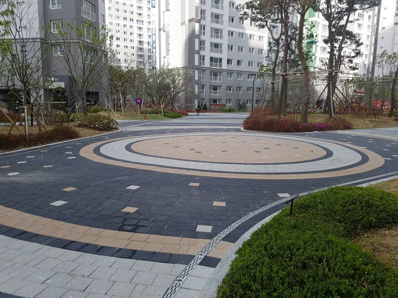 시흥 배곧 한신휴플러스 단지 중앙 원형 광장. 짙은 회색 블록 바탕에 베이지·밝은 회색 블록이 동심원 띠를 이루고 둘레에 화단과 고층 아파트가 있다.