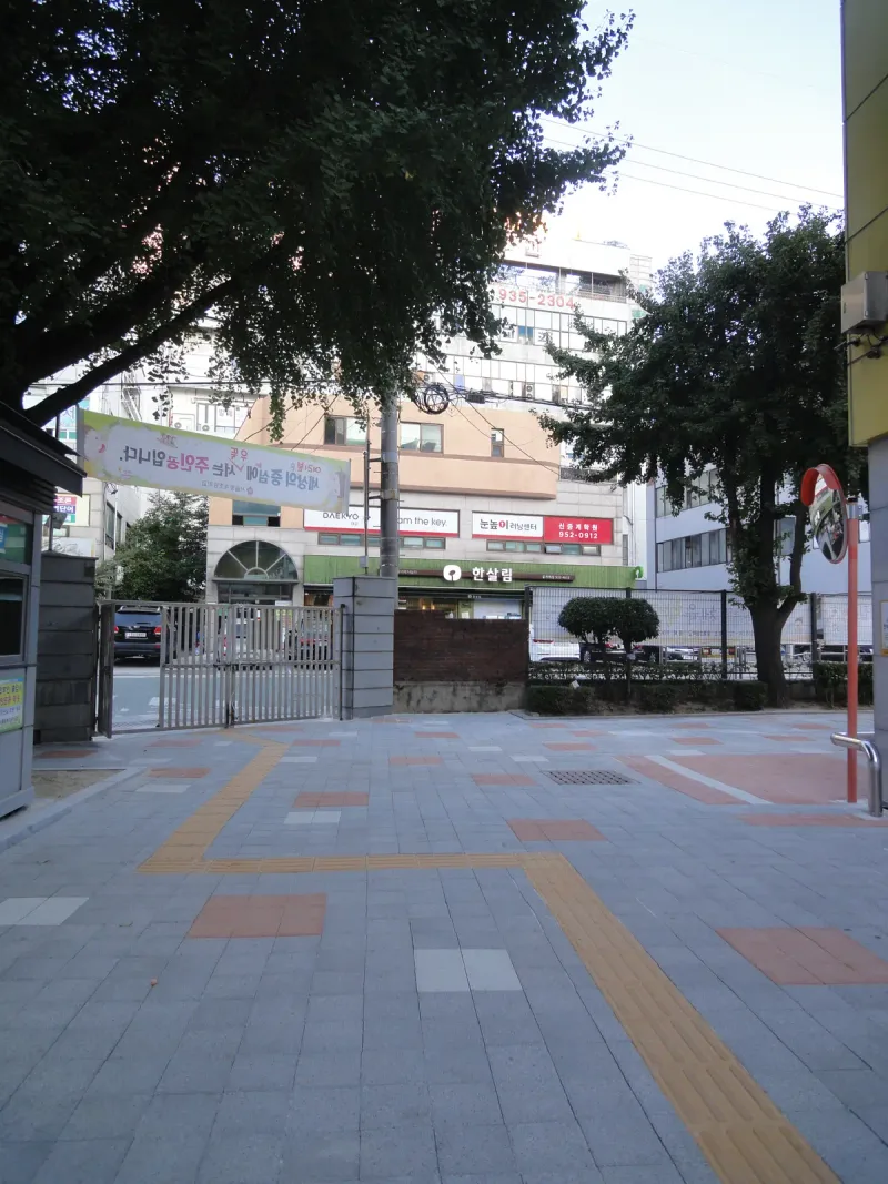 중계초등학교 교문으로 이어지는 바닥. 회색 블록 위로 노란 점자블록이 꺾이며 교문까지 이어지고 적갈색 블록 사각 무늬가 섞여 있다.