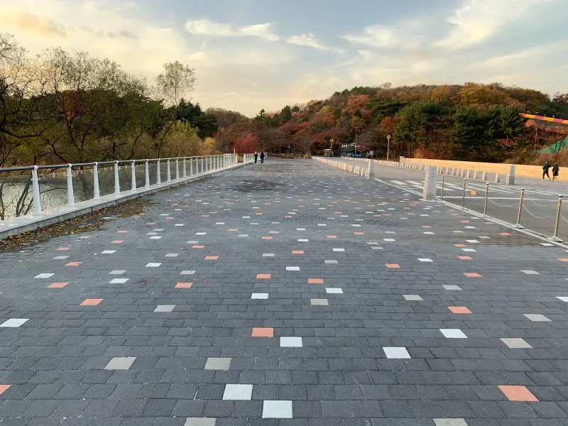 서울대공원 호수 위 넓은 보행로. 흰 난간 사이로 짙은 회색 블록 바탕에 흰색·주황·연회색 블록이 흩뿌려진 길이 산 쪽으로 뻗는다.