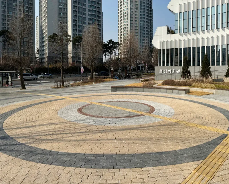 올림픽회관 앞 원형 광장. 회색·베이지·적갈색 블록이 동심원 무늬로 깔려 있고 뒤로 회관 건물과 고층 아파트가 보인다.