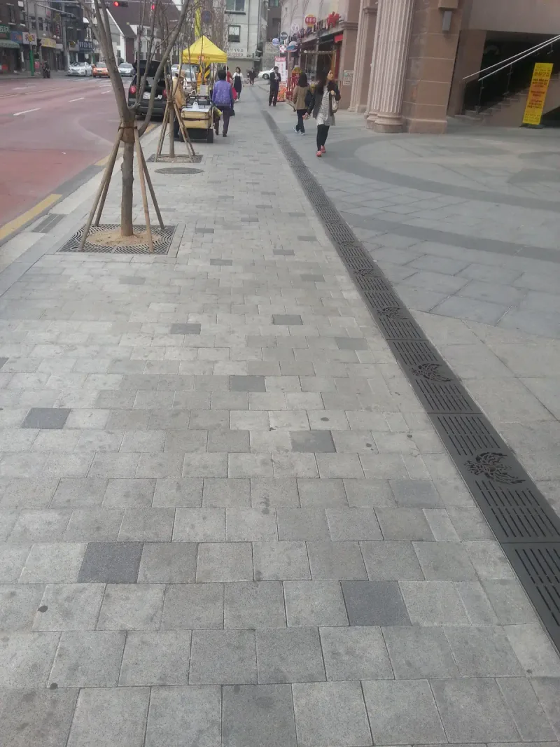 불광 롯데 현장 대로변 보도. 밝은 회색 직사각형 블록에 짙은 회색 블록이 드문드문 섞인 보도 옆으로 가로수 보호판과 무늬 있는 배수 덮개가 길게 이어진다.
