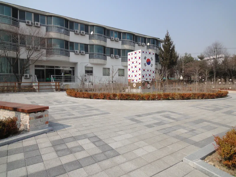 수원보훈원 건물 옆 광장. 번개무늬가 들어간 회색 블록 바닥에 나무 벤치가 놓여 있고 뒤로 건물과 태극기 조형물이 보인다.