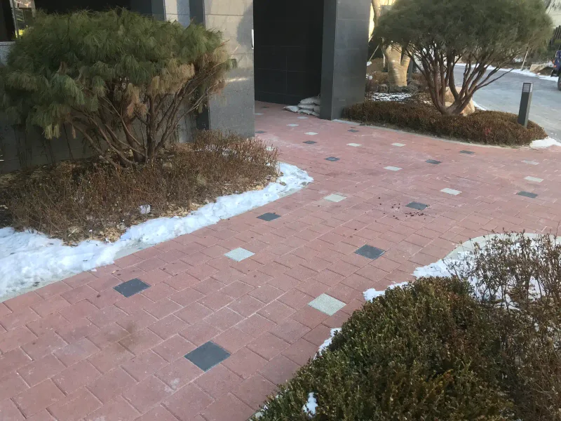 북한산 더샵 화단 모퉁이. 적갈색 블록 길이 소나무와 관목 화단을 끼고 꺾여 들어가며 가장자리에 눈이 남아 있다.