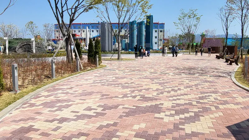 춘천 레고랜드 미니랜드 산책로. 적갈색·분홍·베이지 블록이 지그재그 물결무늬로 깔린 넓은 길이 화단 사이로 뻗고 멀리 블록 모형 건물들이 보인다.