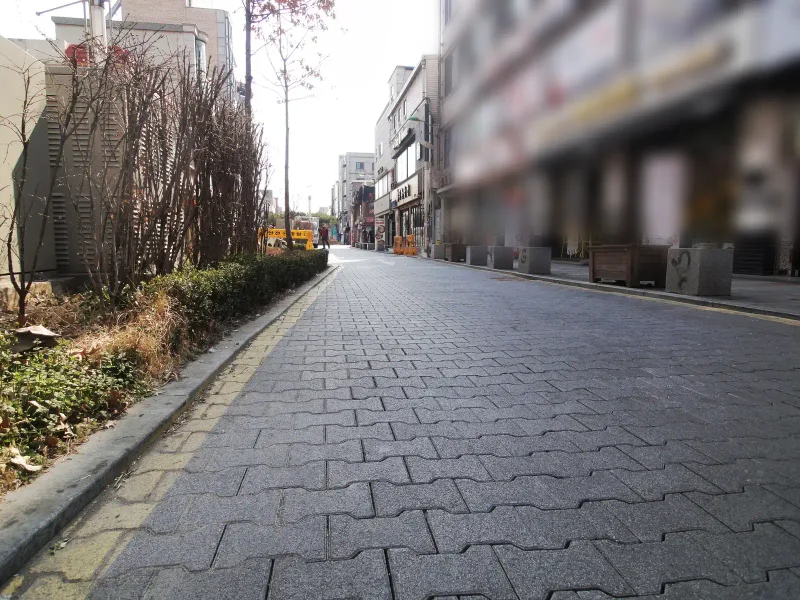 부천대 대학로 상가 거리. 짙은 회색 I형 차도블록이 도로 전체에 깔려 있고 왼쪽에 관목 화단, 오른쪽으로 상가가 이어진다.