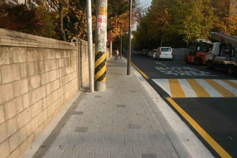 성수문화복지회관 인근 담장 옆 보도. 회색 블록 보도가 블록 담장을 따라 곧게 뻗고, 끝에 노란 횡단보도 표시가 보인다.