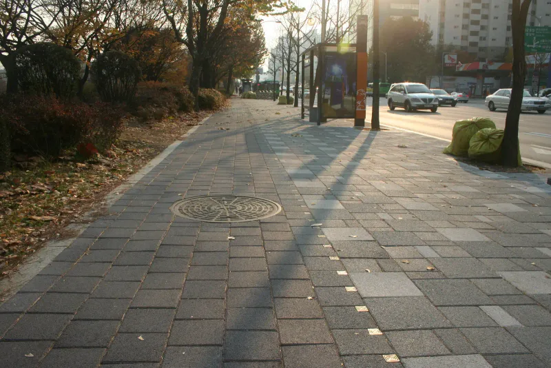 풍납동 버스정류장 옆 보도. 긴 나무 그림자가 드리운 회색 블록 바닥에 맨홀 뚜껑이 있고 밝은 블록이 드문드문 섞여 있다.