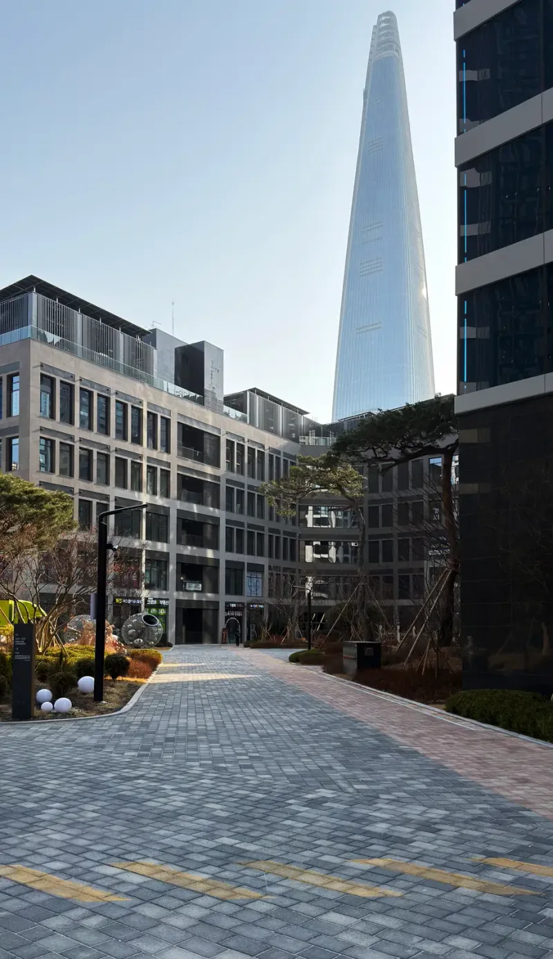 잠실 르엘 단지 가로 풍경. 회색 블록 보행로가 건물 사이로 뻗어 있고 멀리 롯데월드타워가 솟아 있다.
