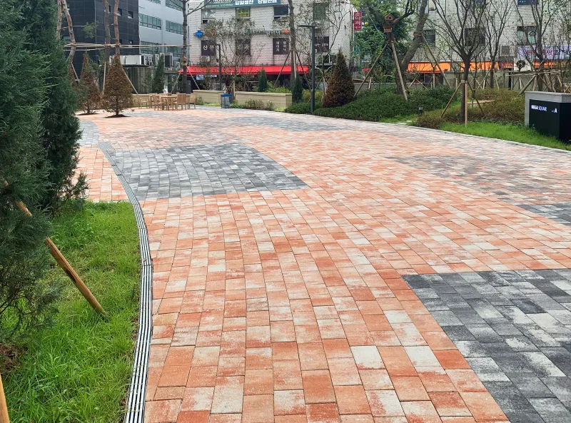 고덕 센트럴 푸르지오 상가 앞 넓은 광장. 주황·적갈색 블록 바탕에 회색 블록 구획이 섞여 있고 곡선 배수로가 화단을 따라 휘어 간다.