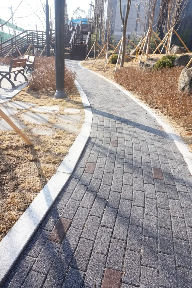 월계 두산위브 벤치 옆 산책로. 짙은 회색 블록 길이 마른 풀밭 사이로 굽어 들어가고 가장자리에 밝은 경계석이 이어진다.