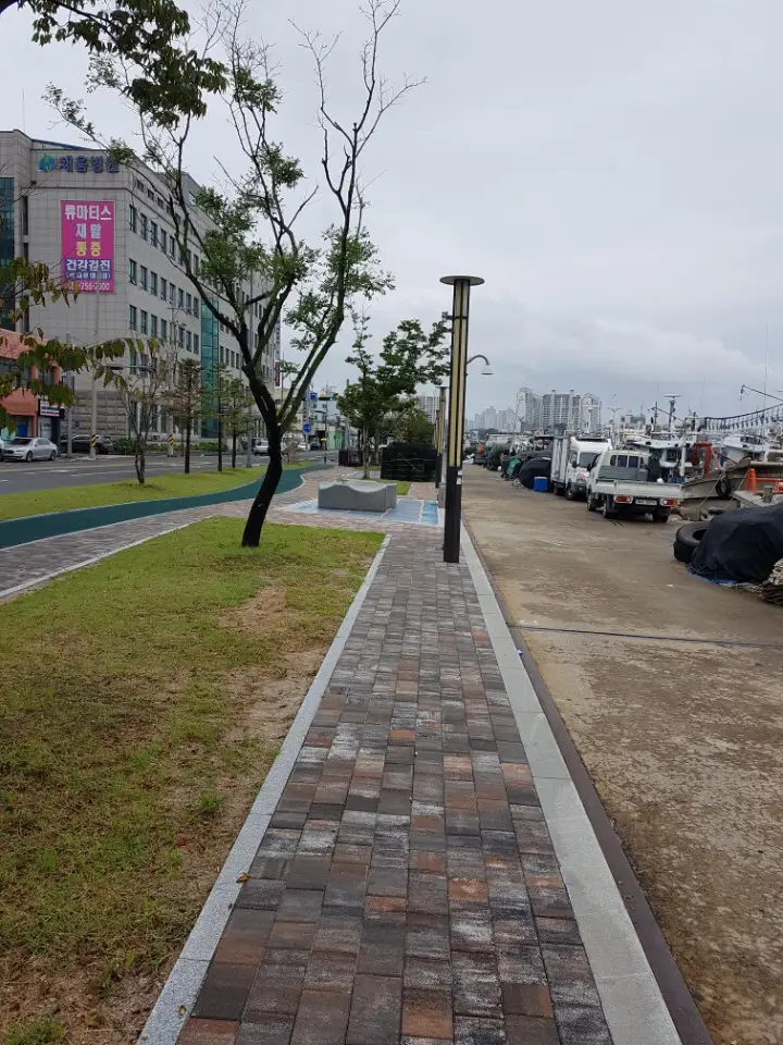 포항 동빈나루 항구 옆 좁은 보행로. 갈색·회색이 섞인 블록 길 한쪽에 잔디 띠가 있고 오른쪽으로 부두와 정박한 배가 보인다.
