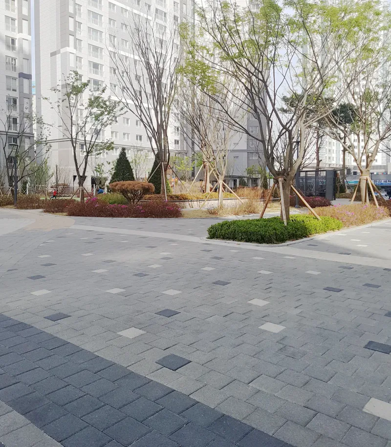 배곧 한신휴플러스 놀이터 옆 광장. 회색 블록 바탕에 진회색과 밝은 블록이 드문드문 박혀 있고 앞쪽으로 진회색 띠가 지나며 둥근 회양목 화단이 보인다.