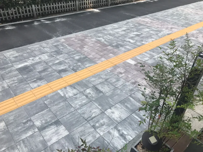 잠실관광특구 보도를 화단 너머로 본 모습. 회색·분홍 계열 블록 위로 노란 점자블록이 비스듬히 지나고 뒤로 흰 울타리 화단이 이어진다.