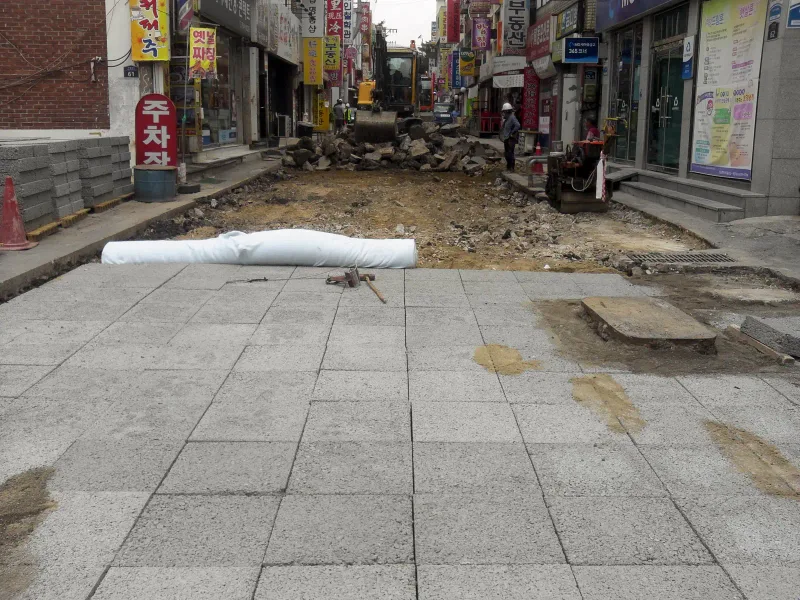 성남 수정로 저작거리의 시공 경계부. 앞쪽은 회색 대형 블록 포장이 끝났고, 그 너머 기존 바닥을 걷어 낸 굴착 구간에 부직포 롤과 굴착기가 보인다.