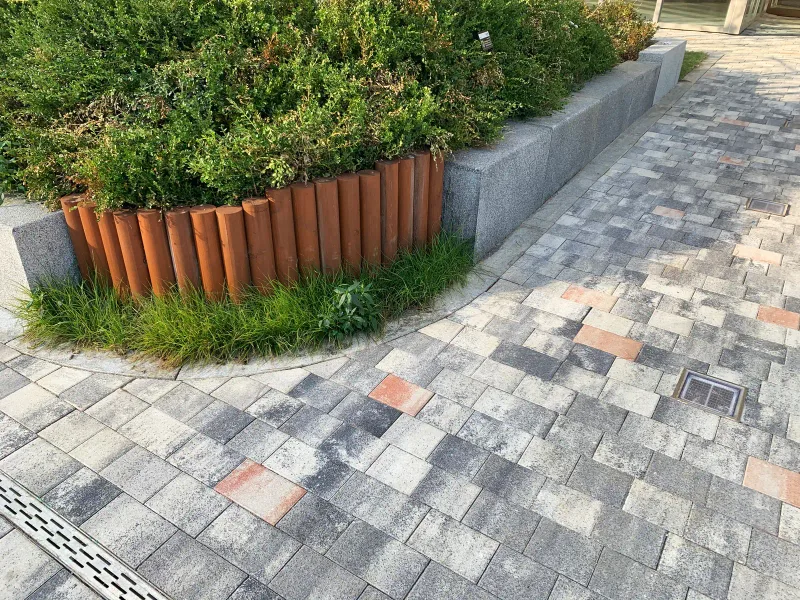 힐스테이트 미사역 12-1BL 곡선 화단 둘레. 원통형 목재 경계와 화강석 경계를 따라 여러 색 블록이 휘어져 깔려 있고 배수 덮개가 보인다.