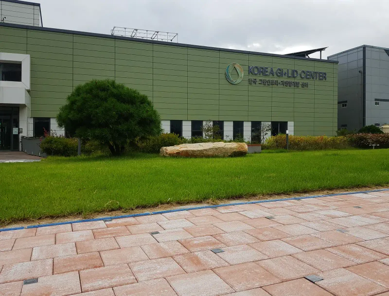 한국 GI·LID 센터 건물 앞. 잔디밭과 소나무 너머로 센터 간판이 달린 건물이 서 있고 앞쪽에 연분홍·베이지 블록 보행로가 지난다.