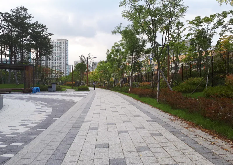 LH 위례택지 단지 옆 산책로. 연회색·회색 블록이 길이 방향 줄무늬로 깔린 길이 가로수와 붉은 관목 화단을 따라 길게 뻗고 왼쪽으로 작은 광장이 이어진다.