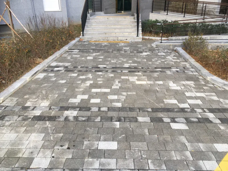 양산 이지더원 2차 동 출입 계단 앞 통로. 회색 블록 바닥에 흰빛·짙은 회색 블록이 흩어져 섞여 있고 짙은 블록 띠가 가로로 두 줄 지나간다.