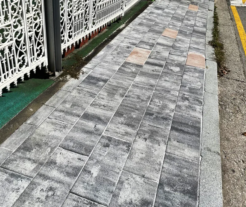 고양 현산초등학교 흰 장식 담장 옆 보도 근접. 회색 결무늬 직사각형 판형 블록과 연분홍 블록이 섞여 있고 화강석 경계석이 차도와 나눈다.