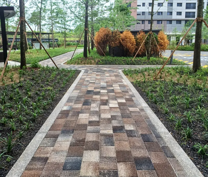 파주 운정 푸르지오 화단 사이 보행로. 짙은 갈색부터 베이지까지 섞인 직사각형 블록이 곧게 깔려 있고 끝에 둥근 관목 화단이 보인다.