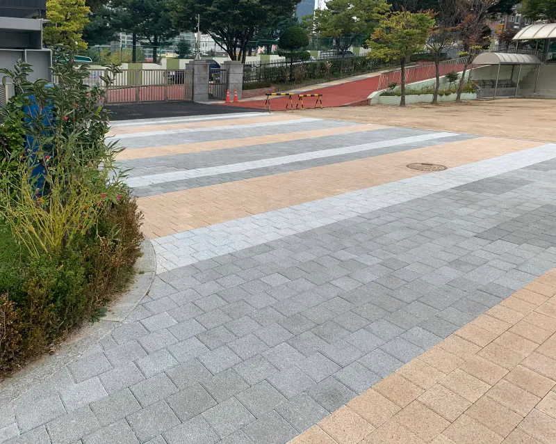 평화초등학교 교문 쪽 진입부. 화단 곡선 경계를 따라 회색·베이지 블록 띠가 깔려 있고 뒤로 붉은 보행로와 담장이 이어진다.
