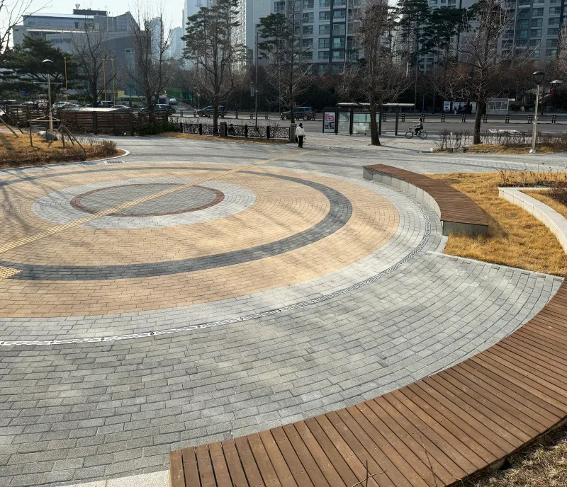 올림픽회관 원형 광장을 옆에서 본 모습. 동심원으로 깔린 베이지·회색 블록 띠를 목재 벤치가 곡선으로 감싼다.