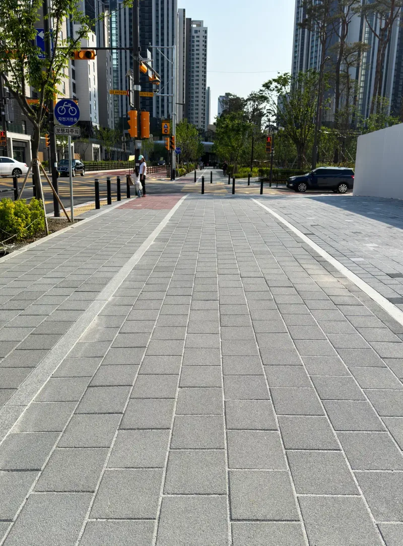 디에이치 퍼스티어 아이파크 교차로 방향 넓은 보도. 짙은 회색 블록 바닥에 흰 경계선이 그어져 있고, 멀리 볼라드와 고층 아파트가 보인다.