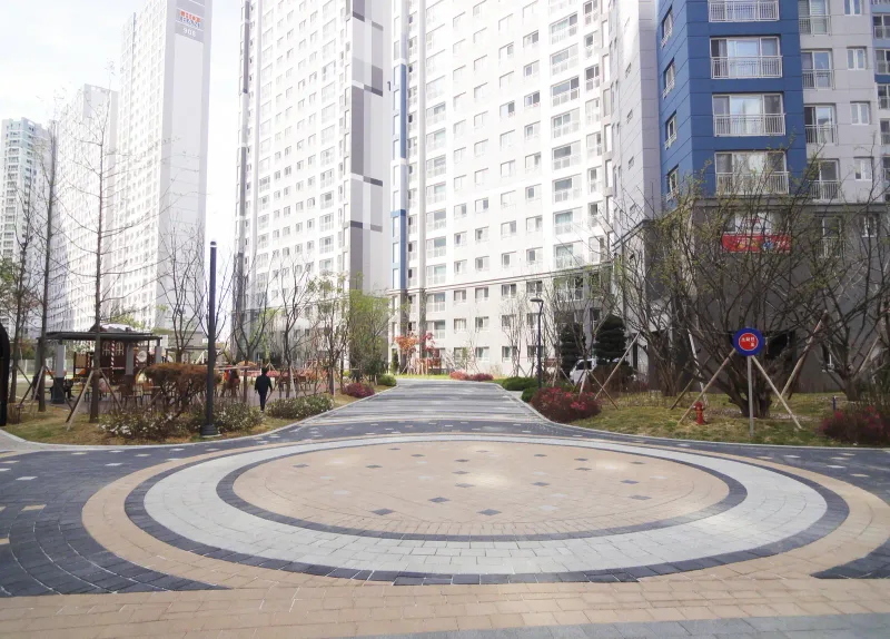 배곧 한신휴플러스 원형 광장을 정면에서 본 모습. 베이지 원 안에 진회색 블록이 마름모꼴로 흩어져 있고 뒤로 놀이터와 동 사이 길이 이어진다.