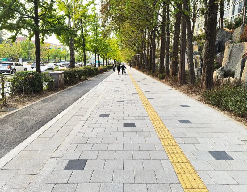 올림픽파크포레온 단지 외곽 가로 보도. 밝은 베이지·회색 판석형 블록 보행로에 노란 점자블록과 짙은 회색 포인트 블록이 박혀 있고 양옆에 가로수가 줄지어 있다.