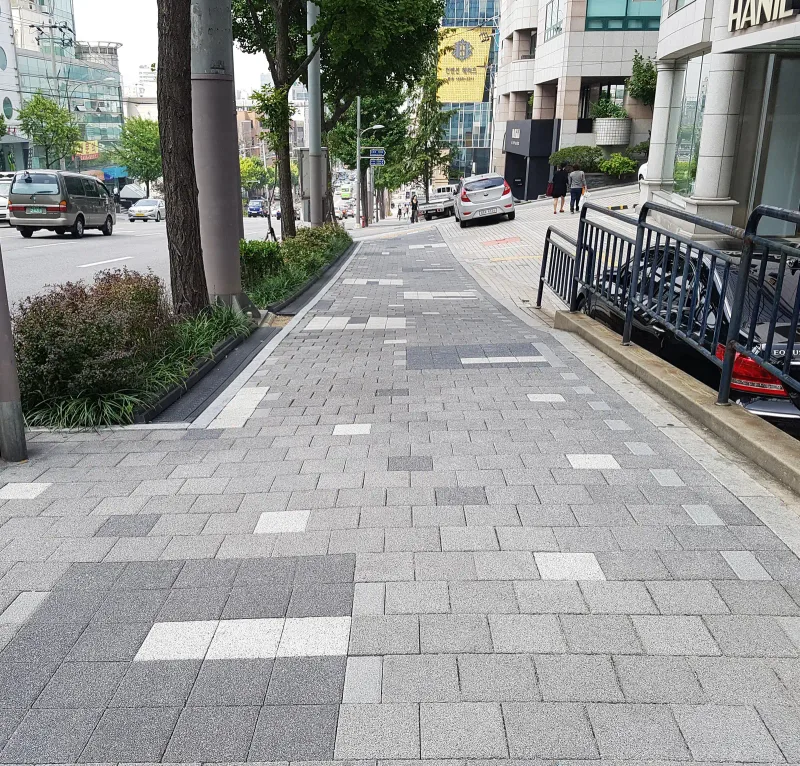 도산대로 오르막 보도. 밝은 회색 직사각형 블록 사이에 짙은 회색 블록 묶음과 흰 블록 줄이 섞여 있고, 왼쪽 화단과 가로수를 따라 보도가 이어진다.