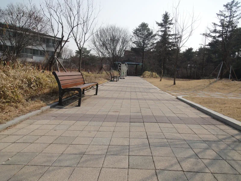 수원보훈원 뒤뜰 산책로. 회색 블록 길 양옆에 나무 벤치가 놓이고 소나무와 겨울나무 사이로 길이 이어진다.