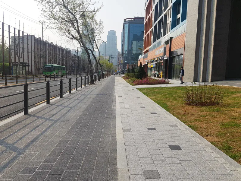개포 래미안 단지 외곽 도로변 보도. 난간 쪽은 짙은 회색 블록, 화단 쪽은 밝은 회색 블록으로 나눠 깔고 멀리 도심 빌딩이 보인다.