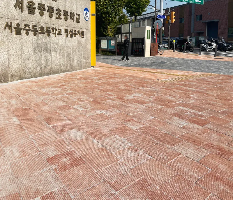 마포 중동초등학교 교명 담장 아래 보도 근접. 분홍·적갈색 줄결 블록이 어긋나게 깔려 있고 담장 끝으로 교문과 도로가 보인다.