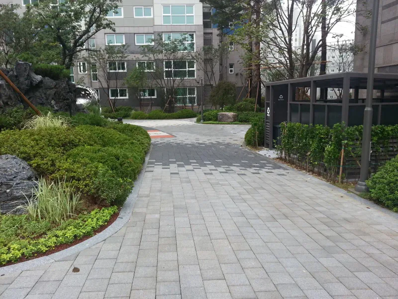 아현동 현장 단지 안 통로. 회색 블록 길이 조경석과 관목 화단 사이로 휘어져 아파트 동 앞으로 이어진다.