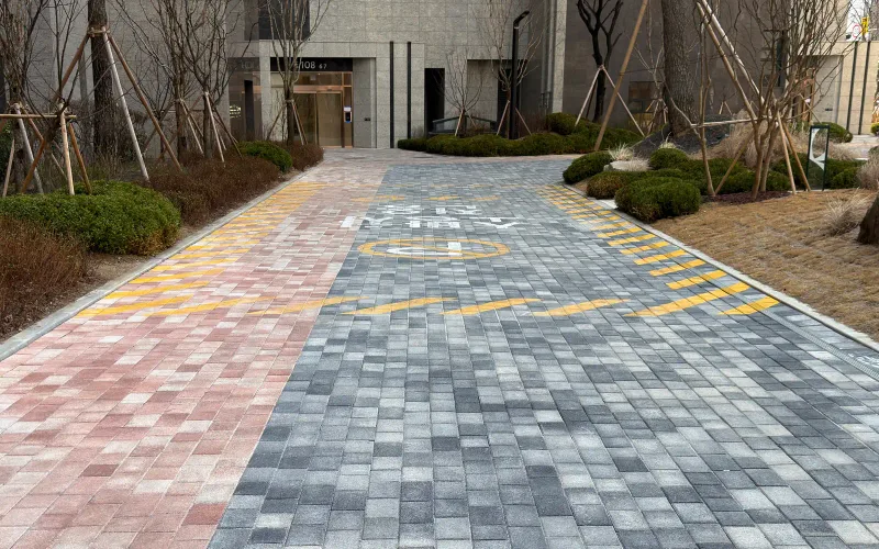 잠실 르엘 동 출입구 앞 진입로. 분홍 계열 블록과 회색 계열 블록이 반씩 나뉘어 깔리고 노란 사선 도색이 더해져 있다.