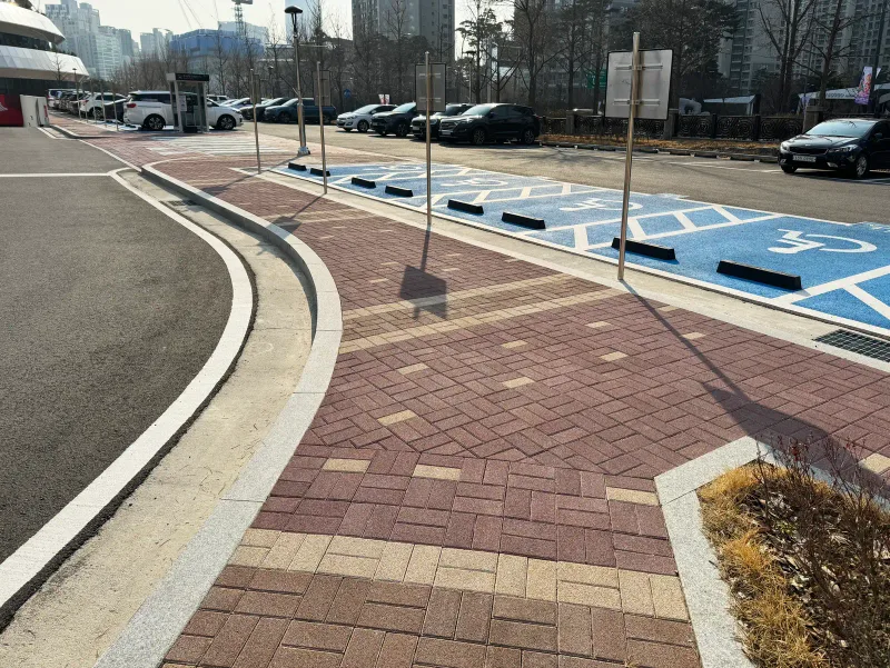 올림픽회관 주차장 옆 곡선 보도. 적갈색과 베이지 블록이 짜임 무늬로 깔리고 옆에 파란 장애인 주차면이 있다.