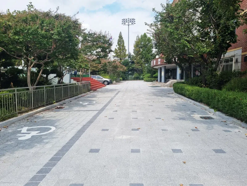 중원초등학교 교내 진입로. 밝은 회색 블록 바닥에 짙은 회색 블록 한 줄이 길게 이어지고 장애인 주차면 표시가 그려져 있다.