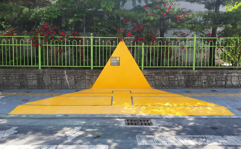 옐로카펫블록 설치 현장, 학교 울타리 앞 보도. 노란 삼각형 벽판 아래 바닥이 노란색으로 덮여 있고 가운데와 앞쪽에 노란 점자블록 줄이 깔려 있다.