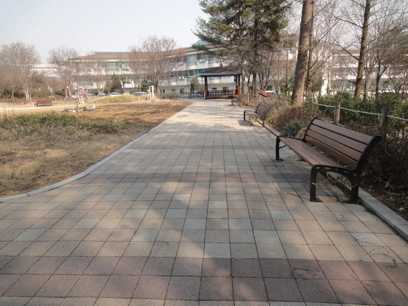 수원보훈원 정원 산책로. 연회색 블록 길 옆에 나무 벤치가 있고 멀리 정자와 건물이 보인다.
