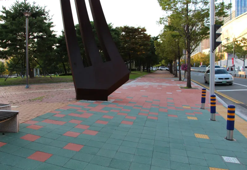 부산 영화의전당 조형물 주변. 청록색과 적갈색 블록이 섞인 넓은 보행 공간이 가로수 길 쪽으로 이어진다.