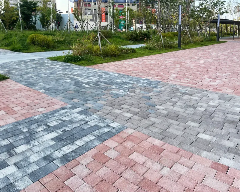 다산자이 아이비플레이스 광장 바닥. 적갈색 블록 면과 회색·흰색이 섞인 블록 띠가 엇갈려 만나며 뒤로 화단과 나무가 이어진다.