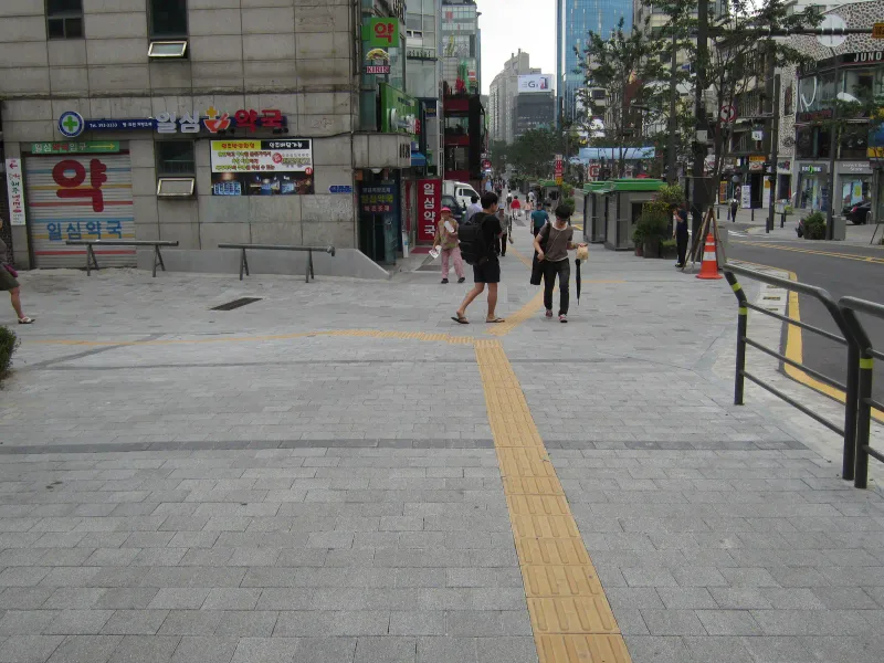 신촌 연세로 교차 지점. 회색 블록 바닥에서 노란 점자블록이 두 갈래로 나뉘고 짙은 회색 블록 띠가 가로지르며, 오른쪽에 보행 난간이 서 있다.
