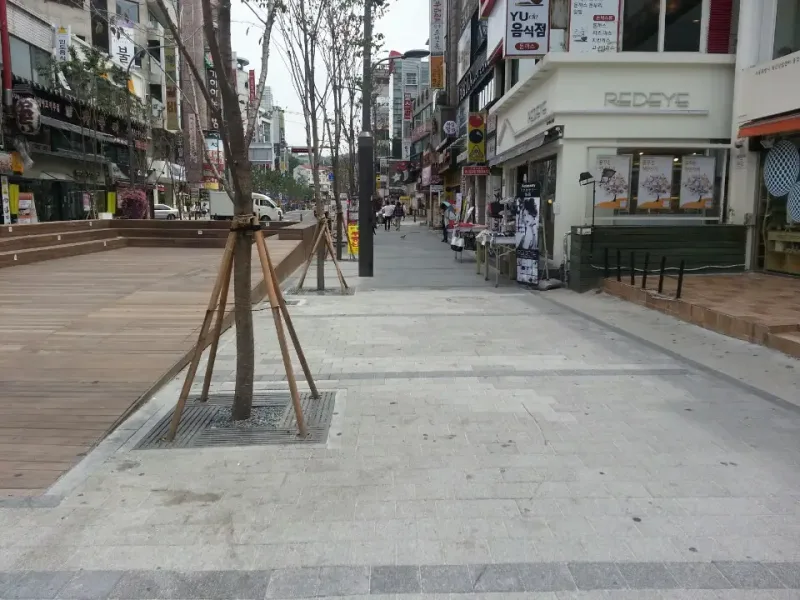 신촌 연세로 가로수 주변. 회색 블록 보도에 새로 심은 가로수와 보호판이 놓이고, 옆으로 나무 데크 쉼터가 이어진다.