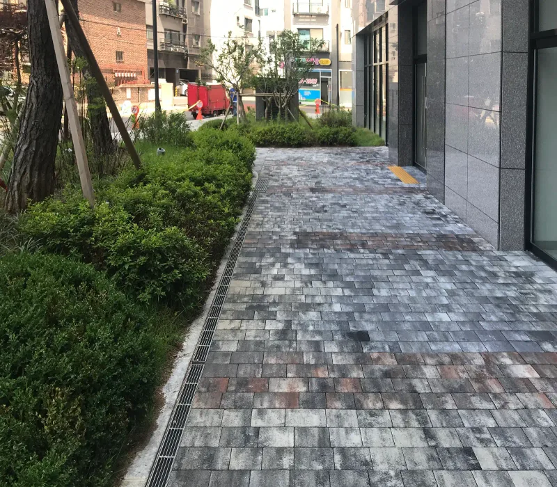 자양 래미안 프리미어 팰리스 상가 옆 보도. 배수 덮개와 관목 화단을 따라 짙은 회색·흰색·적갈색 블록이 섞인 길이 건물을 끼고 이어진다.
