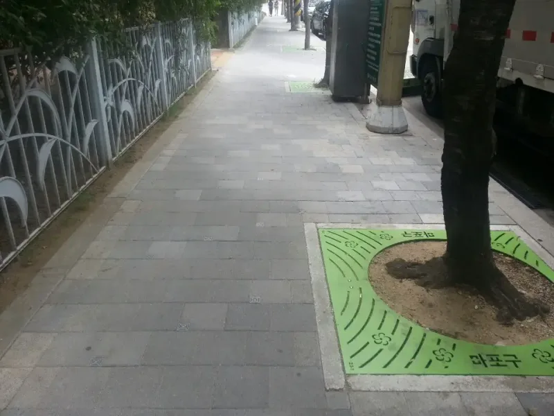 합정 배수분구 주택가 보도. 회색 블록 보도가 흰 철제 울타리를 따라 길게 이어지고, 연두색 가로수 보호판을 두른 나무가 서 있다.