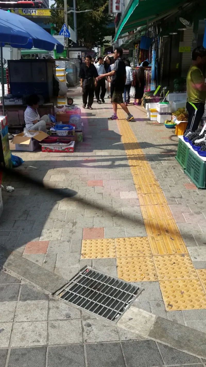 남대문시장 노점 앞 보도 모퉁이. 연회색 블록에 적색·흰 블록이 점점이 섞인 바닥 위로 노란 점자블록이 이어지고 앞쪽에 빗물받이 덮개가 있다.